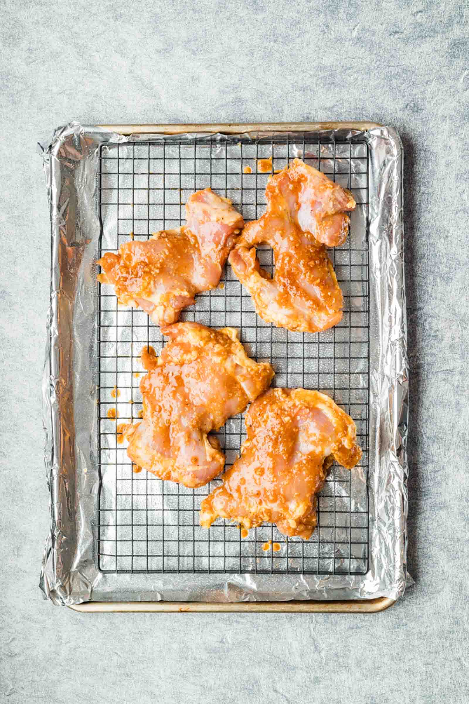 Four pieces of marinated raw chicken on a wire rack over a foil-lined baking sheet.