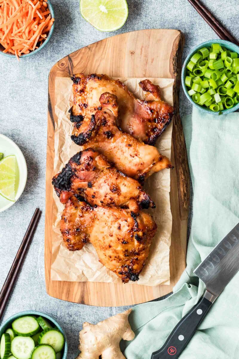 Lemongrass chicken pieces on parchment paper atop a wooden board, surrounded by sliced cucumbers, lime, shredded carrots, green onions, and a kitchen knife.