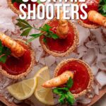 A plate of shrimp cocktail shooters in glasses with spiced rims, filled with cocktail sauce, garnished with parsley and lemon wedges, served on ice.