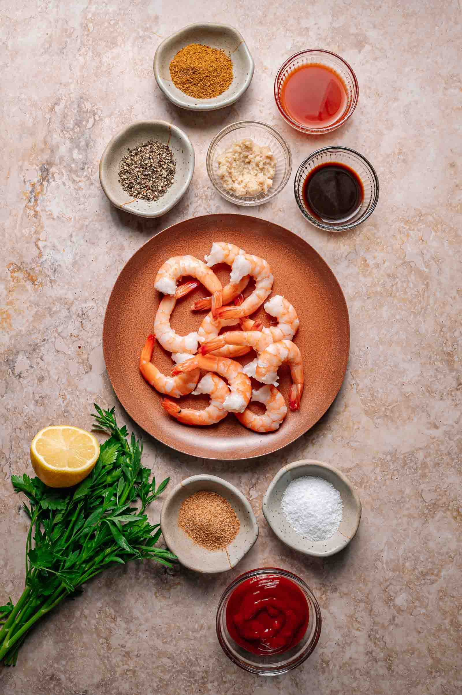 A plate of cooked shrimp is surrounded by small bowls of spices, sauces, minced garlic, lemon, parsley, salt, pepper, and ketchup on a light stone surface.
