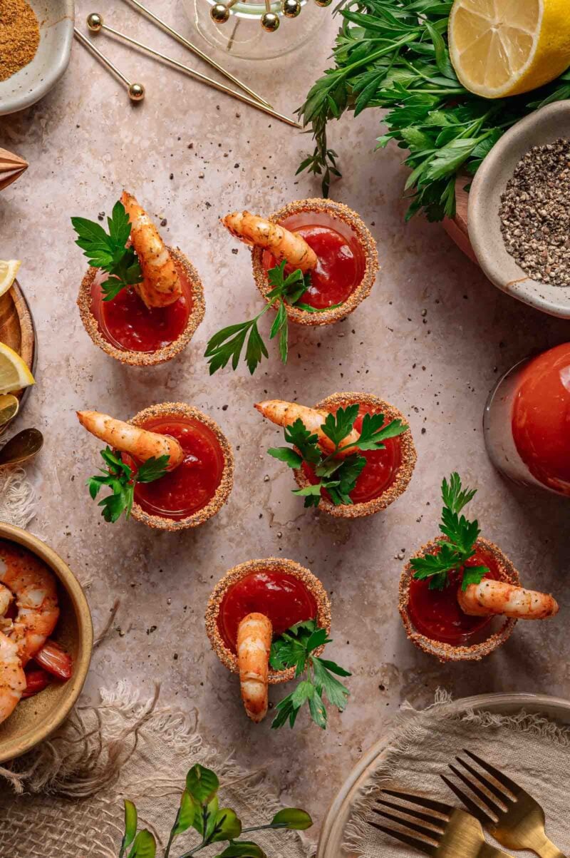 Six small cups filled with cocktail sauce, each garnished with a shrimp and parsley, are arranged on a textured surface, surrounded by herbs, lemon, pepper, and utensils.