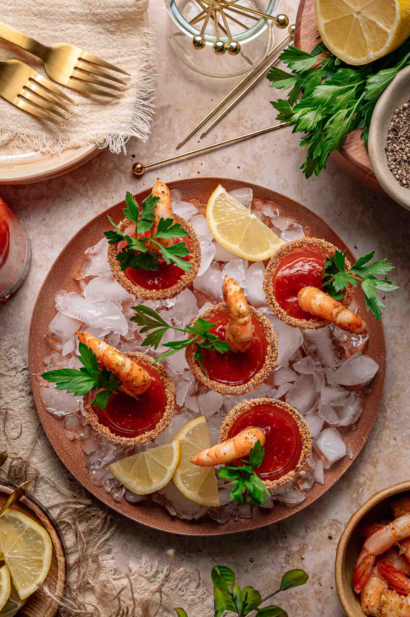 A plate of shrimp cocktail shots served in glasses rimmed with seasoning, garnished with parsley and lemon wedges, placed on ice.