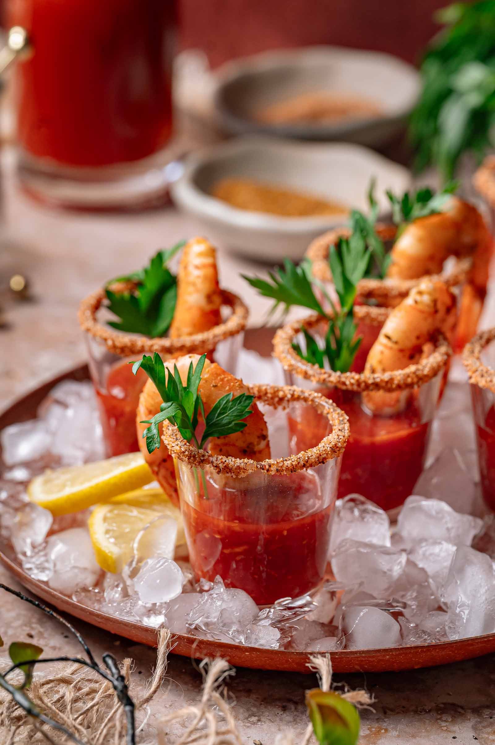 Shrimp cocktail shooters in glasses rimmed with seasoning, garnished with parsley and lemon slices, served on ice.