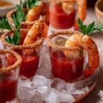 Close-up of shrimp cocktail shooters in glasses rimmed with seasoning, garnished with parsley, and served on a bed of ice.