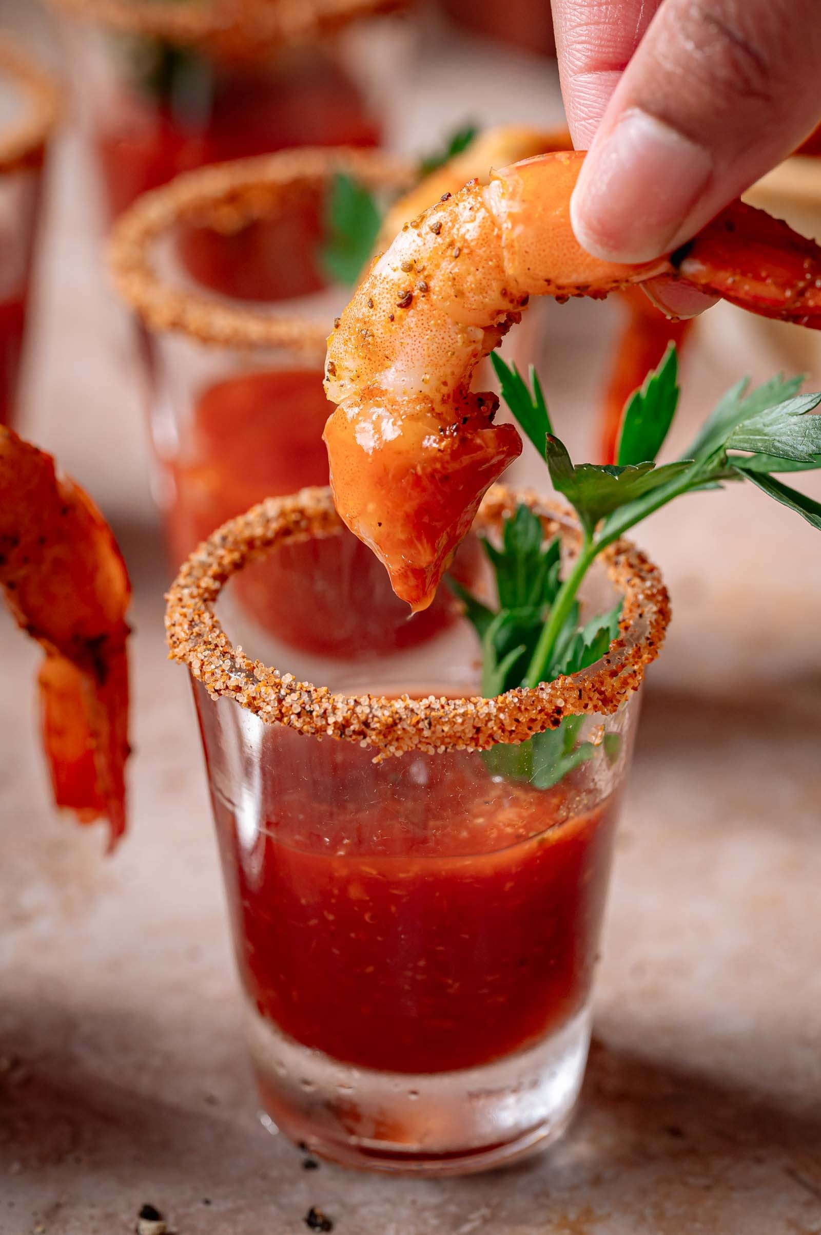 A hand dips a seasoned shrimp into a shot glass filled with cocktail sauce, with a parsley garnish and a spiced rim.