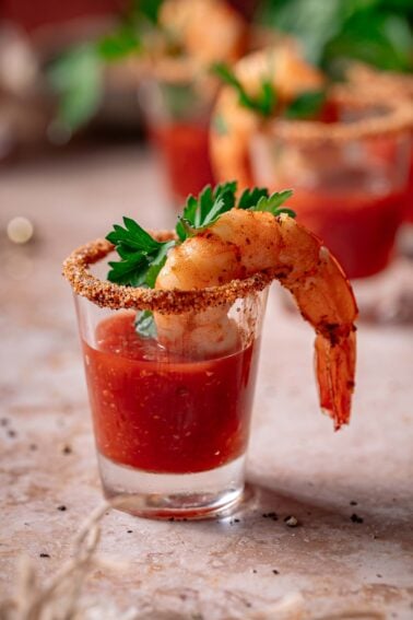 A shot glass filled with cocktail sauce, garnished with a shrimp, a parsley leaf, and a seasoned rim.