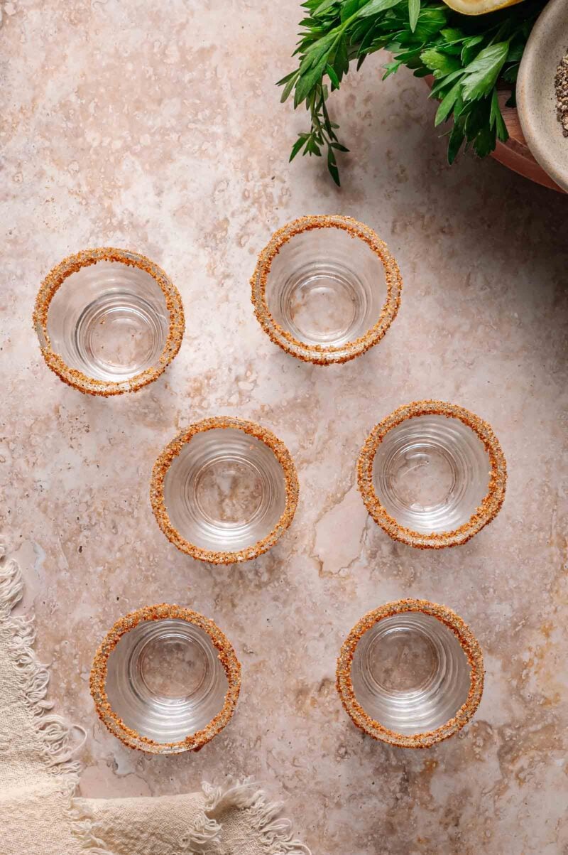 Six empty glasses with seasoned rims are arranged on a marble surface, with fresh herbs and a beige textured cloth visible at the edges.