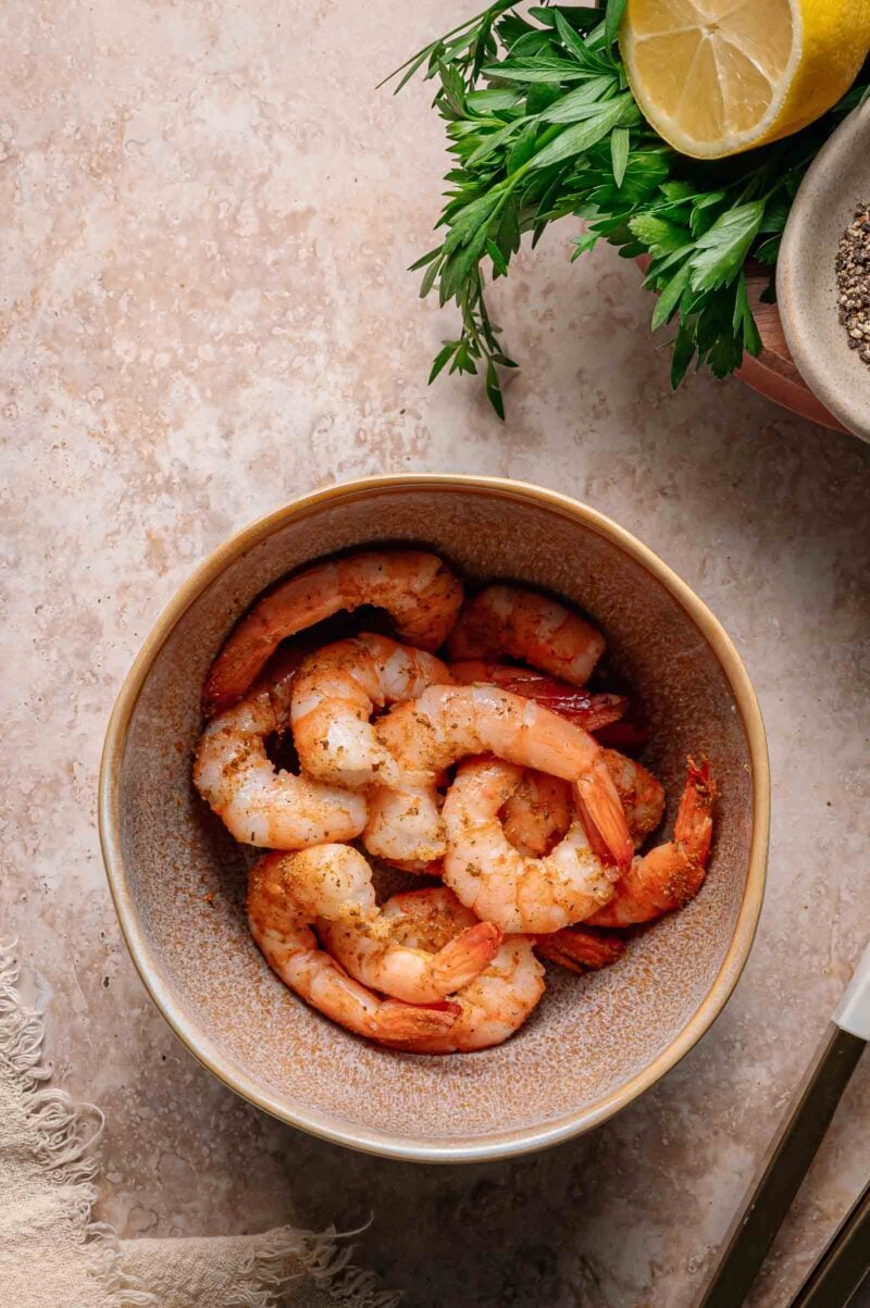 A bowl of cooked, seasoned shrimp sits on a stone surface next to fresh parsley, half a lemon, and a bowl of ground black pepper.