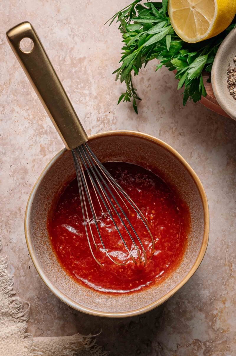 A bowl of red cocktail sauce with a metal whisk, next to fresh parsley, half a lemon, ground pepper, and a textured cloth on a beige surface.