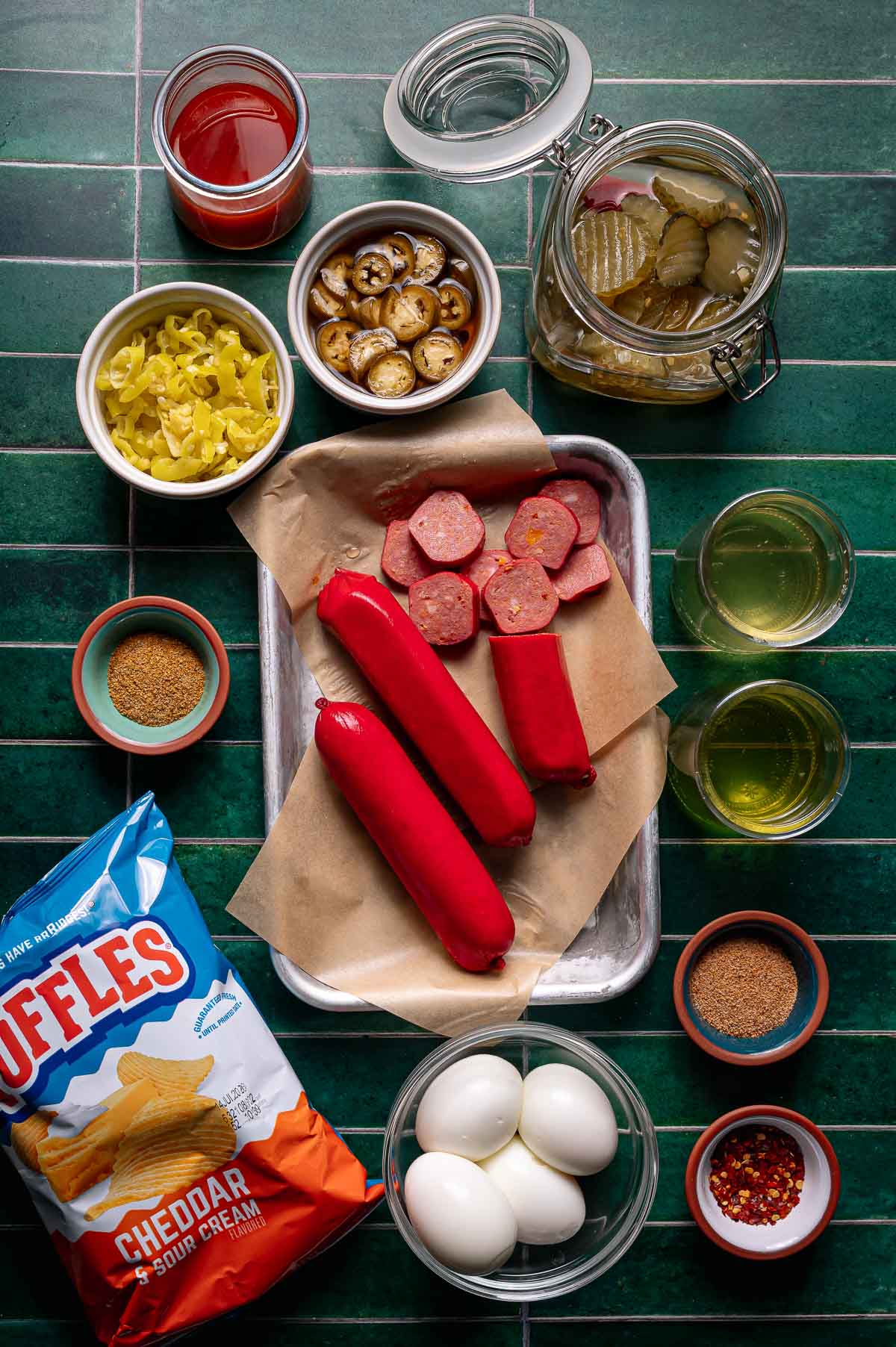 A flat lay of the viral spicy bowl ingredients with red sausages, hard-boiled eggs, chips, pickles, peppers, spices, and jars on a green tiled surface.