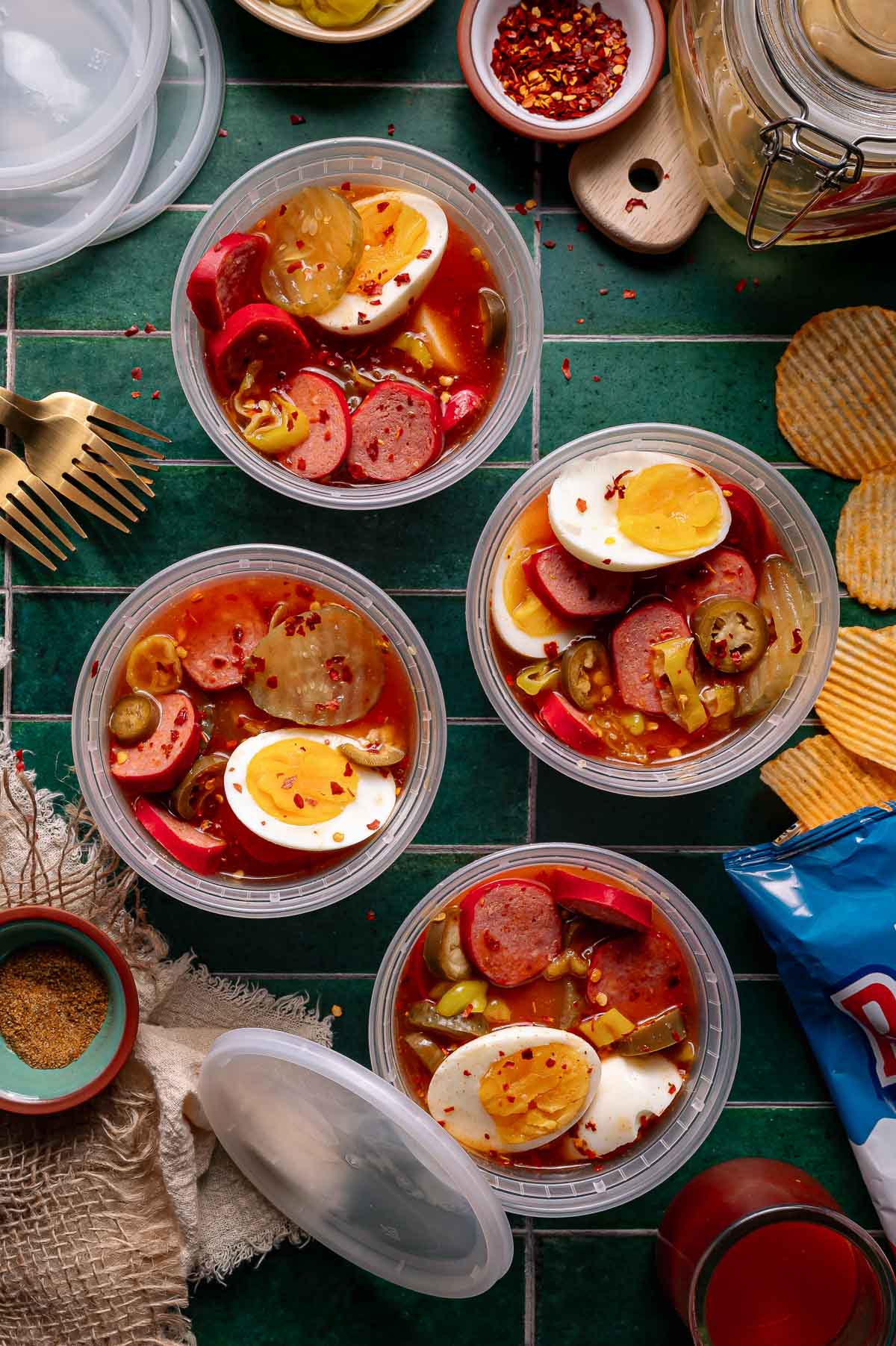 Four plastic containers filled with eggs, sausage slices, jalapeños, and pickled vegetables in a spicy red liquid, arranged on a green tiled surface with crackers and utensils nearby.