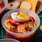 A plastic container filled with sliced sausage, hard-boiled egg, jalapeños, onions, and spicy brine, with potato chips in the background.
