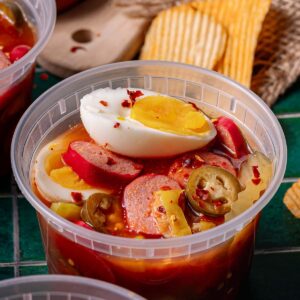 A plastic container filled with sliced sausage, hard-boiled egg, jalapeños, onions, and spicy brine, with potato chips in the background.