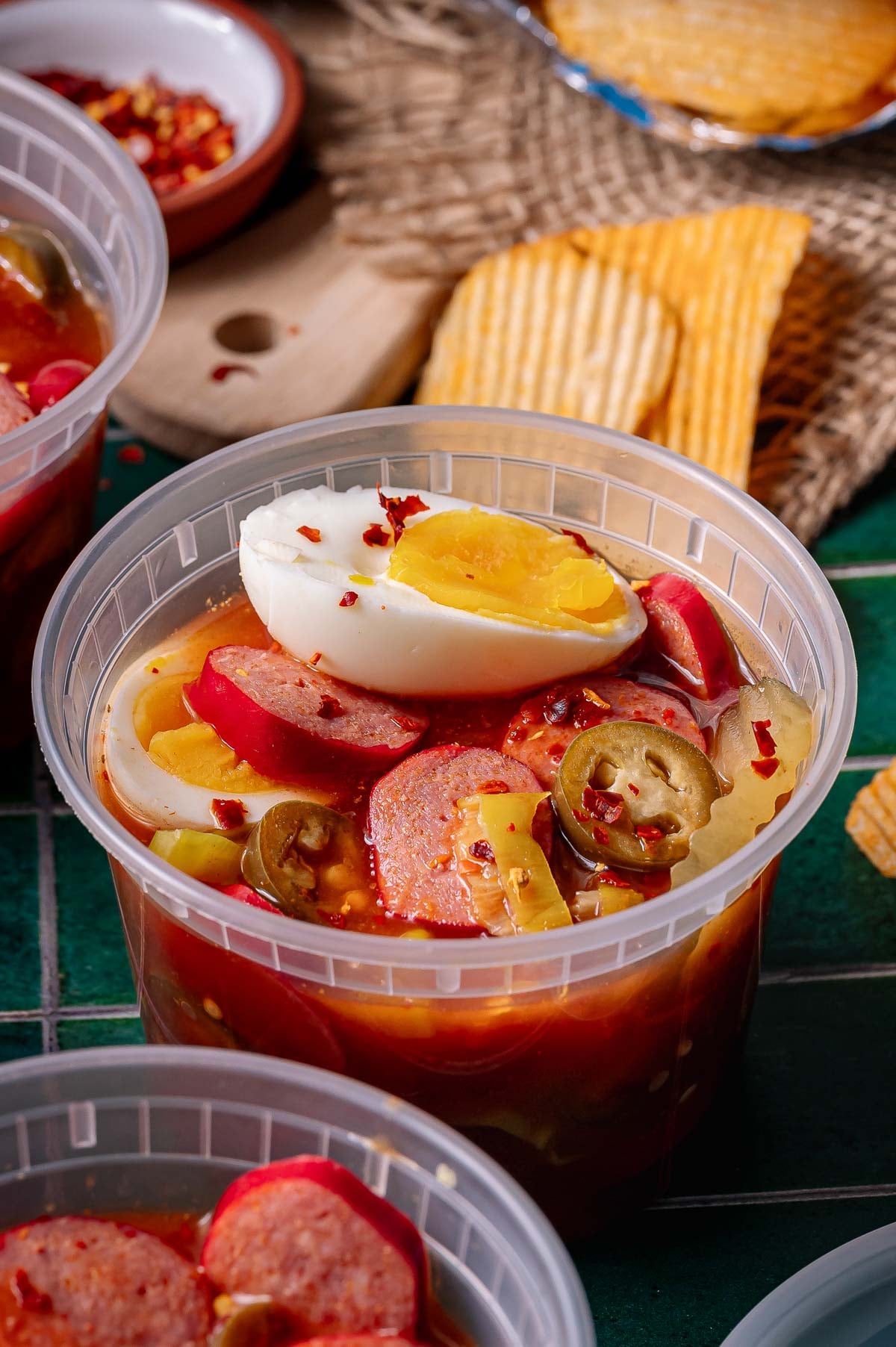 A plastic container filled with sliced sausage, hard-boiled egg, jalapeños, onions, and spicy brine, with potato chips in the background.