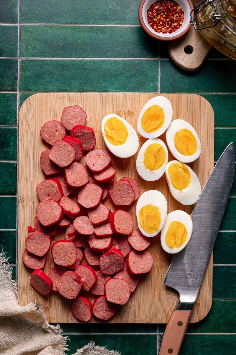 Sliced sausage and halved hard-boiled eggs arranged on a wooden cutting board with a knife on a green tiled surface.