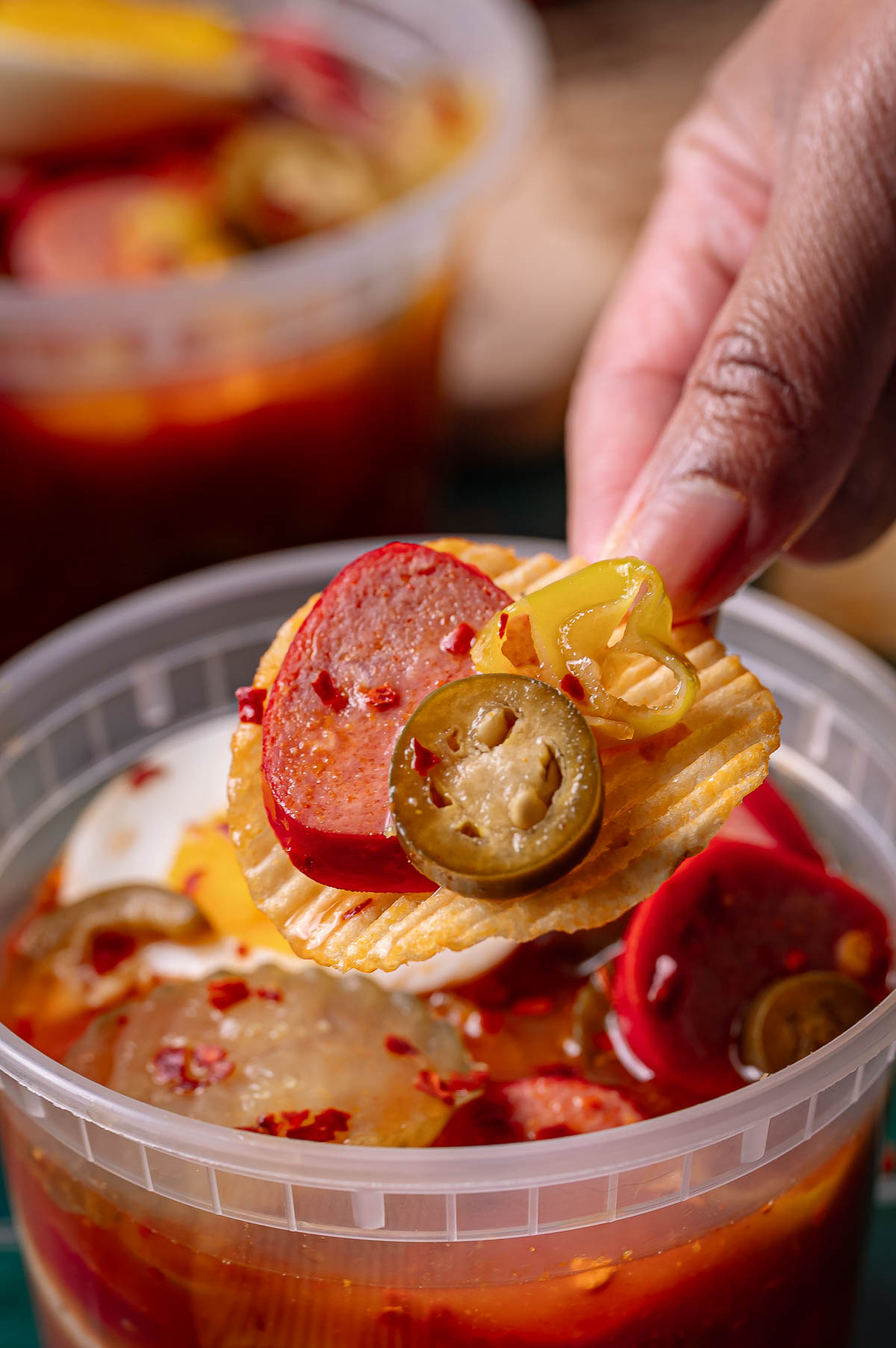 A hand holds a ridged potato chip topped with sausage slices and pickled jalapeños over a container of red, spicy bowl.
