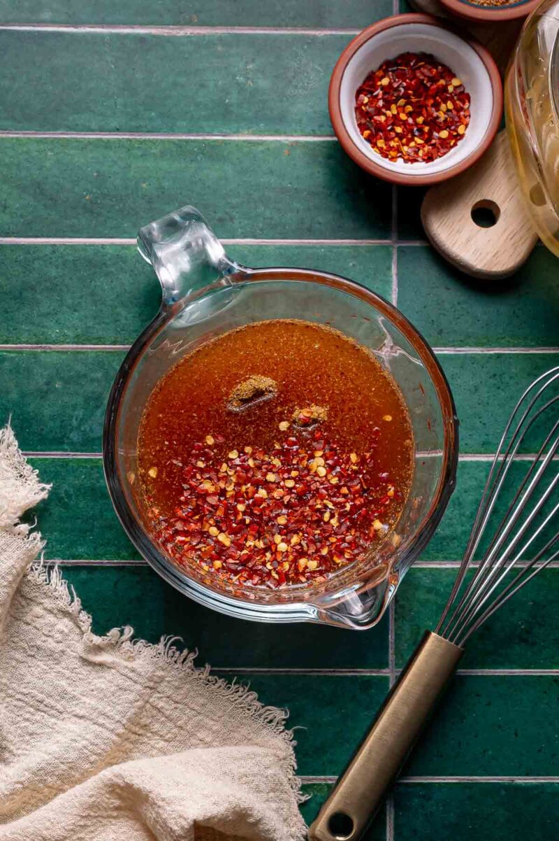 A glass measuring cup containing a reddish-brown liquid with chili flakes, next to a whisk, a small bowl of chili flakes, and a beige cloth on a green tiled surface.