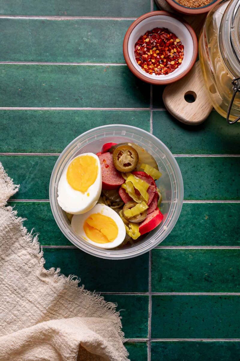 A plastic container with two halved boiled eggs, pickled jalapeños, and sliced vegetables on a green tiled surface near a beige cloth and bowls of spices.