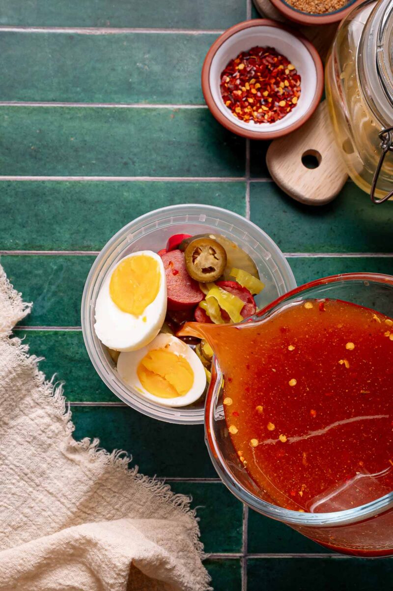 A plastic container with hard-boiled eggs, sliced sausage, and pickled jalapeños next to a bowl of red liquid on a green tiled surface.