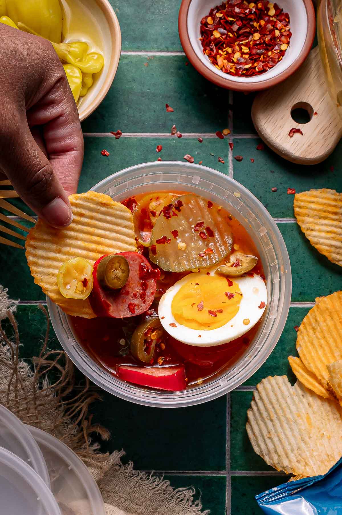 A hand dips a ridged potato chip into a spicy bowl with and hard-boiled egg slices, with more chips and seasonings on a green tiled surface.