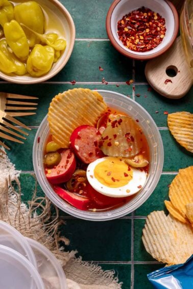 The viral spicy bowl with sliced pickles, boiled eggs, red sausage, banana peppers, and potato chips, topped with red pepper flakes on a green tile surface.