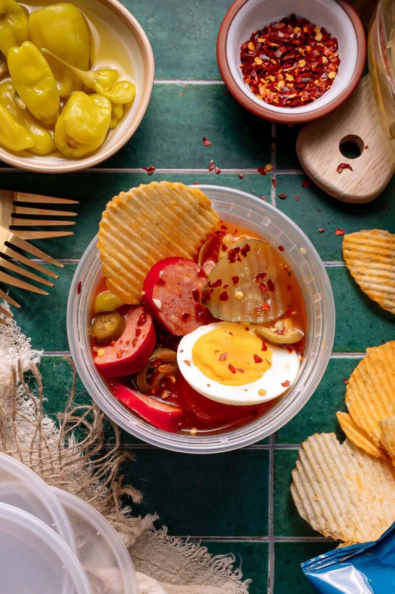 The viral spicy bowl with sliced pickles, boiled eggs, red sausage, banana peppers, and potato chips, topped with red pepper flakes on a green tile surface.