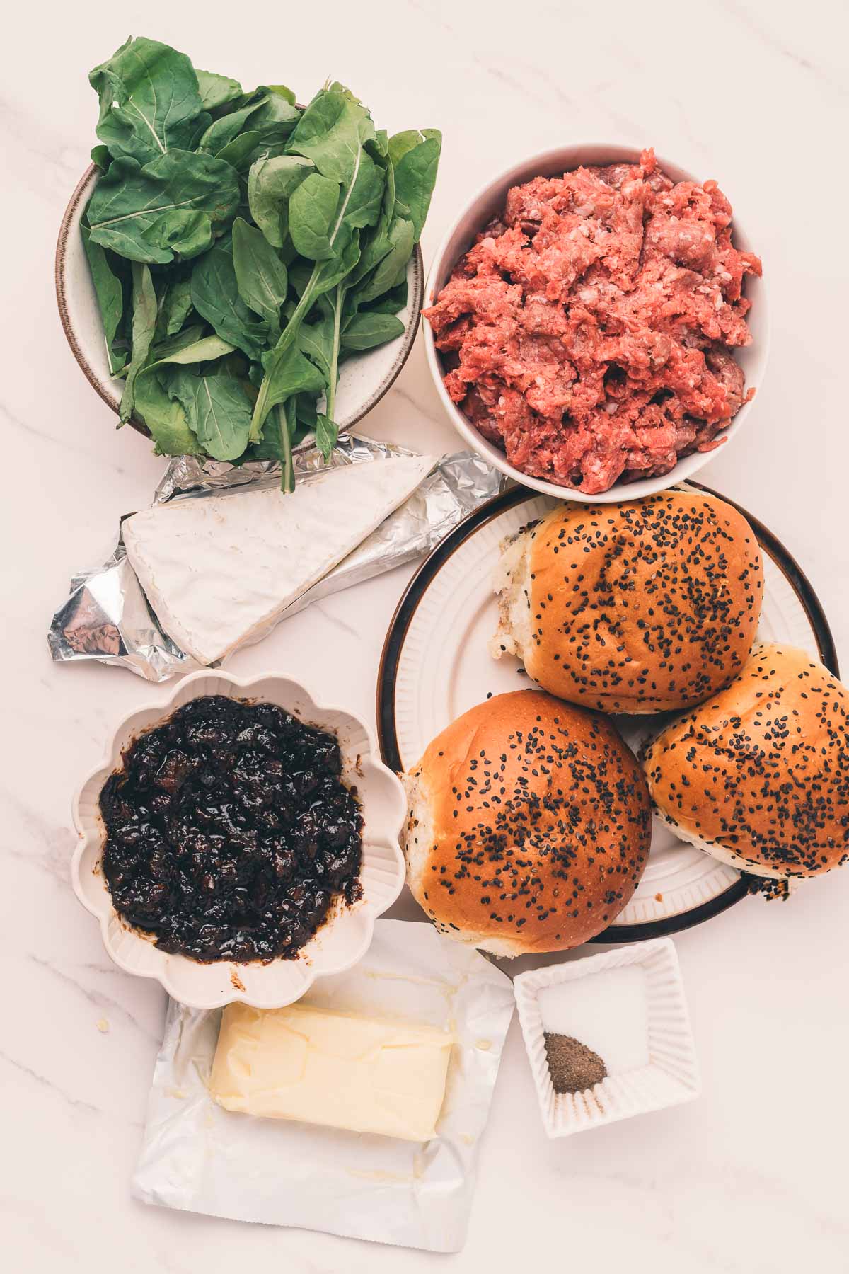 Top-down view of burger ingredients: ground beef, leafy greens, brie cheese, butter, seed-topped buns, bacon jam, and a small dish of black pepper.