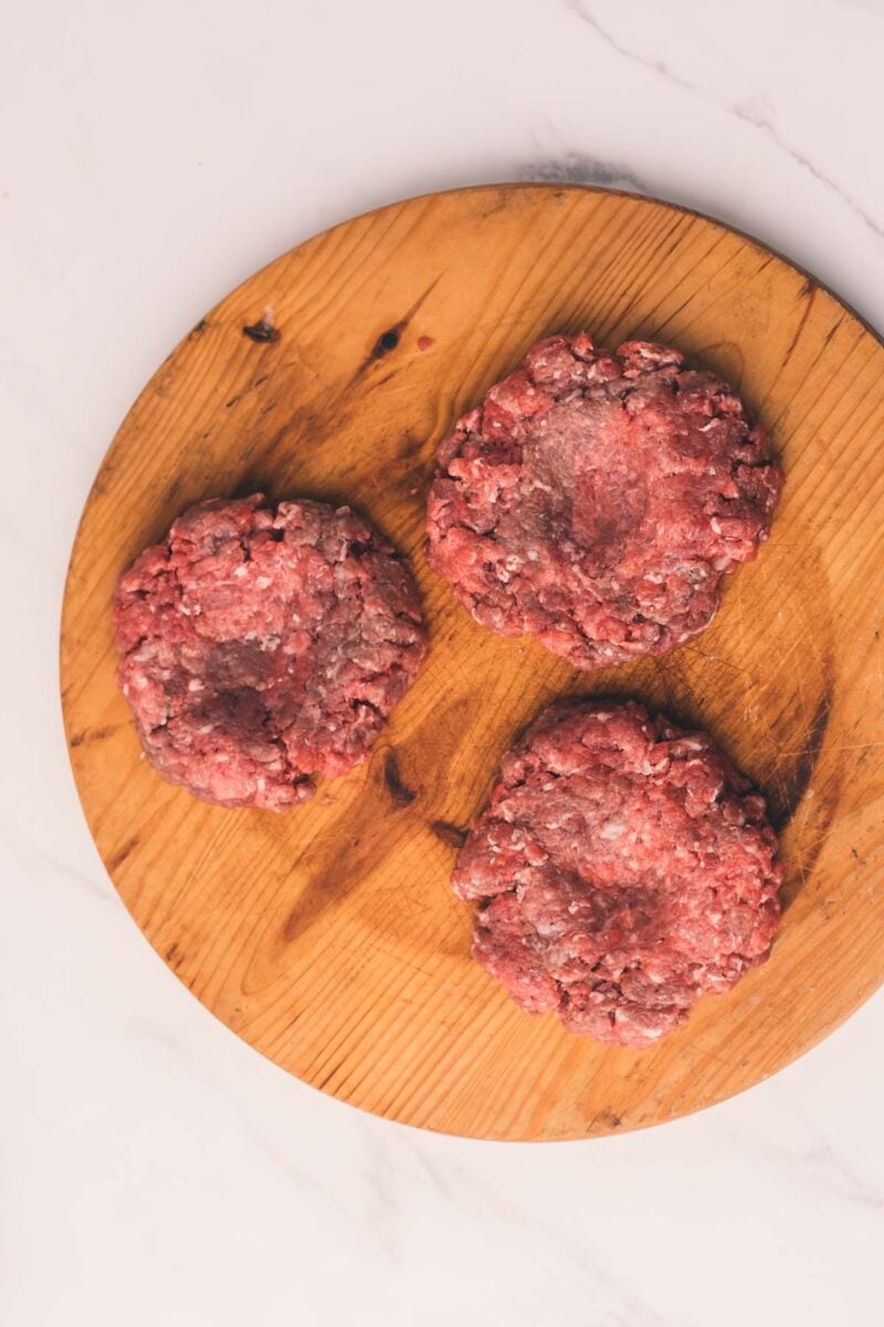 Three raw ground beef patties on a round wooden cutting board, viewed from above on a light-colored surface.