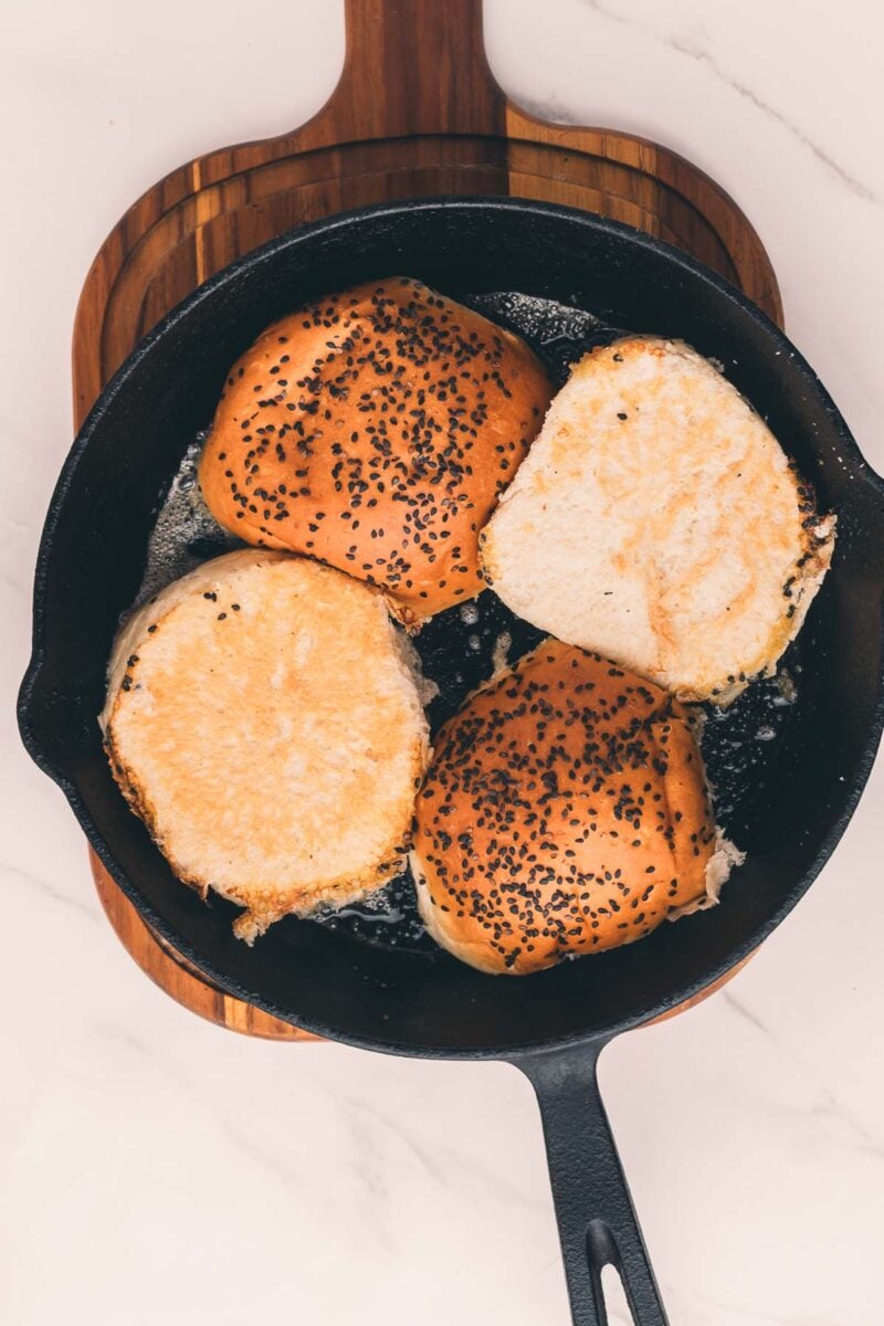 Four burgers buns are toasting cut-side down in a black cast iron skillet on a wooden cutting board.
