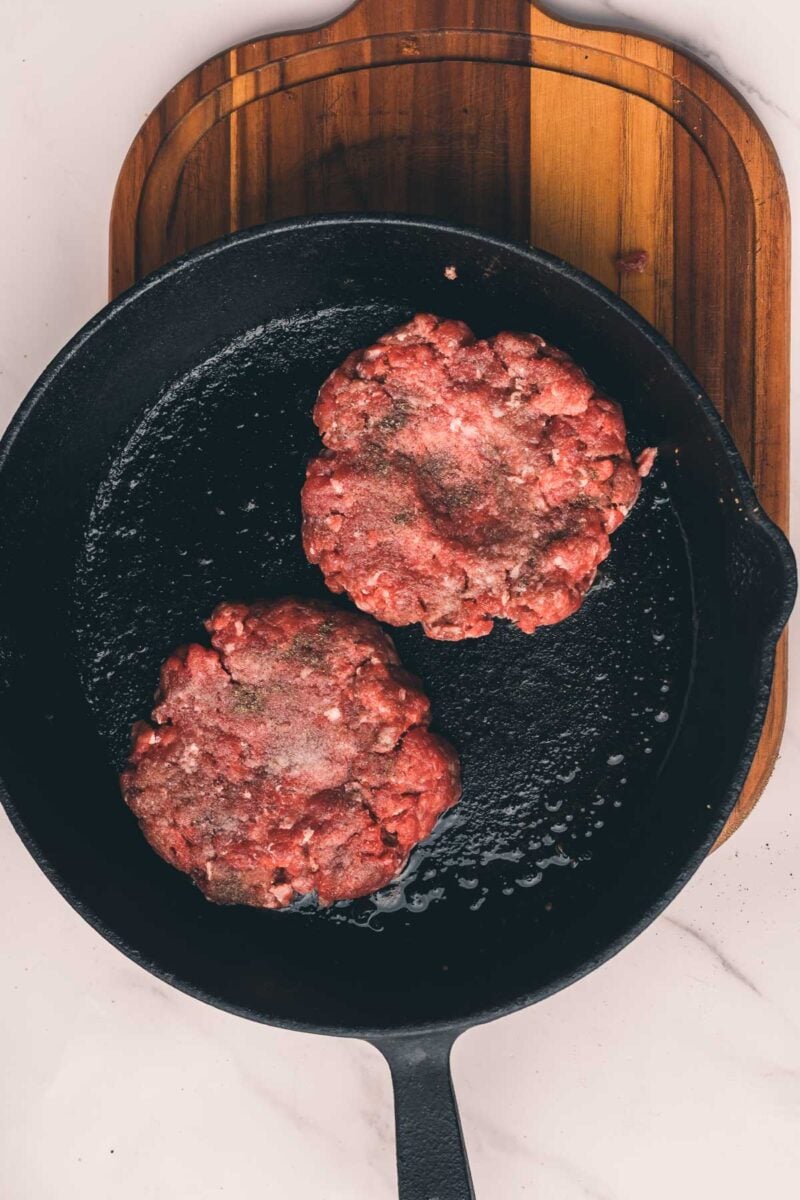 Two raw beef burger patties seasoned with pepper in a black cast iron skillet on a wooden cutting board.