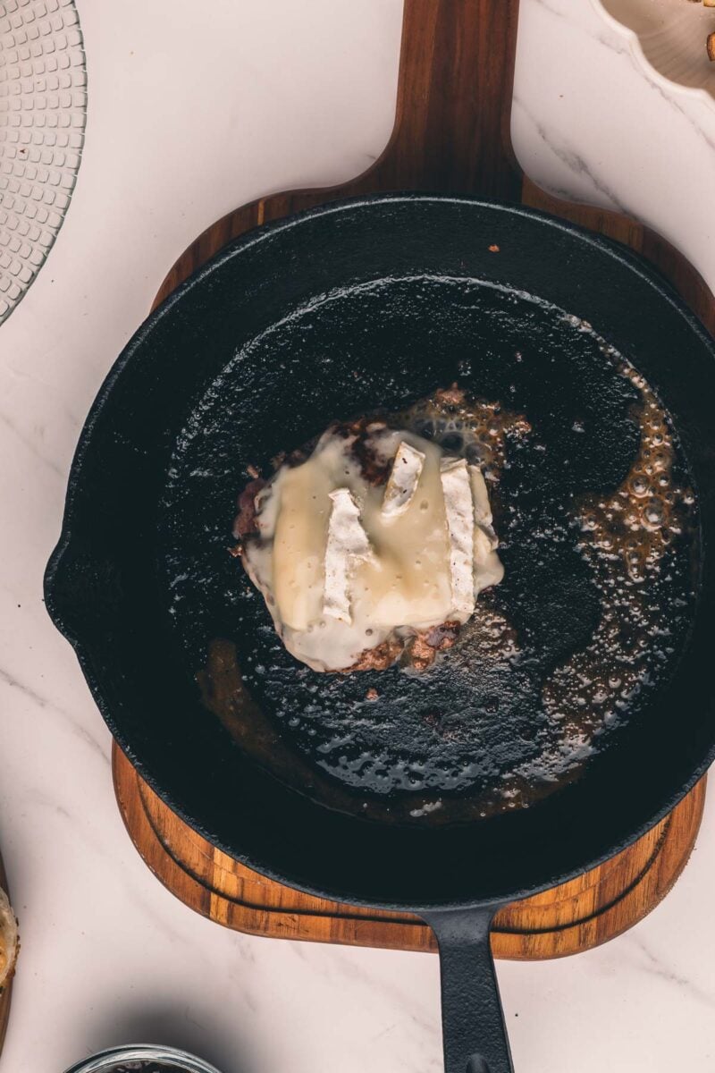 A hamburger patty topped with melting brie is cooking in a black cast iron skillet on a wooden trivet.