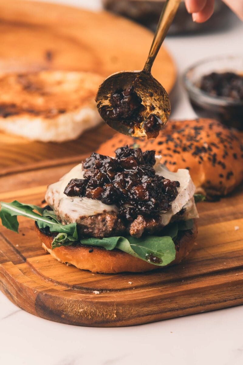 A spoon is adding bacon jam onto a cheeseburger with greens on a wooden board; a seeded bun and more chutney are in the background.