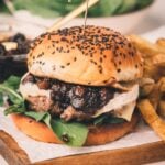 A burger with leafy greens, cheese, and bacon jam in a sesame seed bun, served with fries on a wooden board.