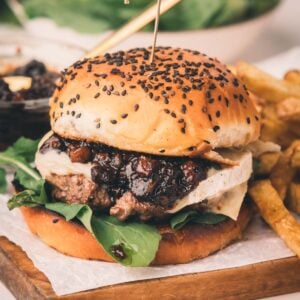 A burger with leafy greens, cheese, and bacon jam in a sesame seed bun, served with fries on a wooden board.