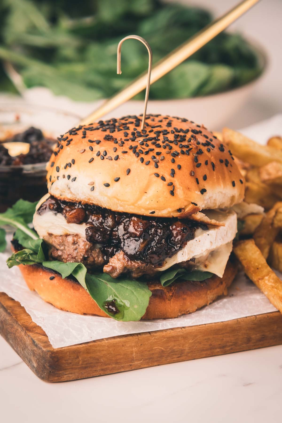 A burger with leafy greens, cheese, and bacon jam in a sesame seed bun, served with fries on a wooden board.