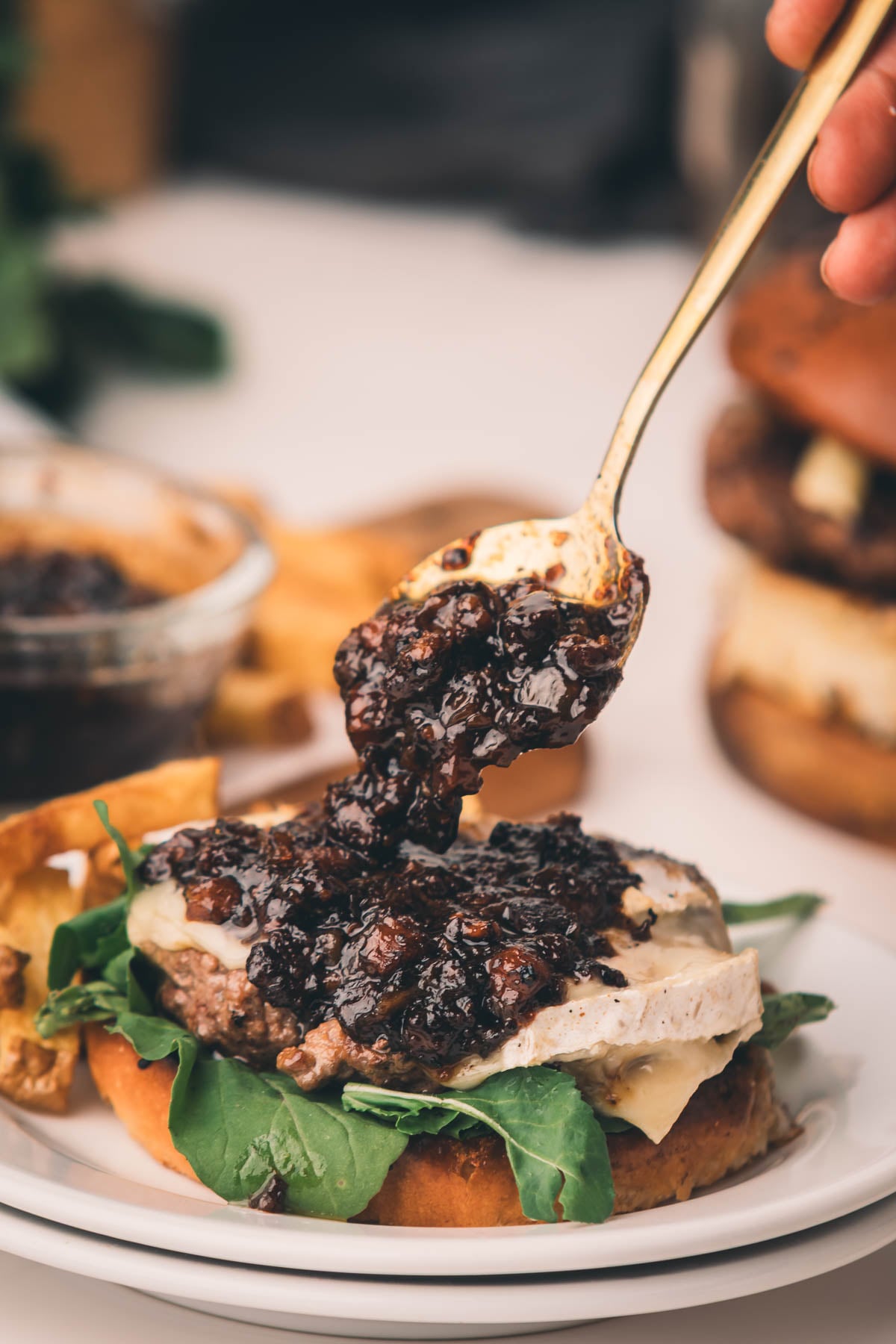 A hand spoons dark bacon jam onto a cheeseburger with greens on a plate, with chips and another burger in the background.