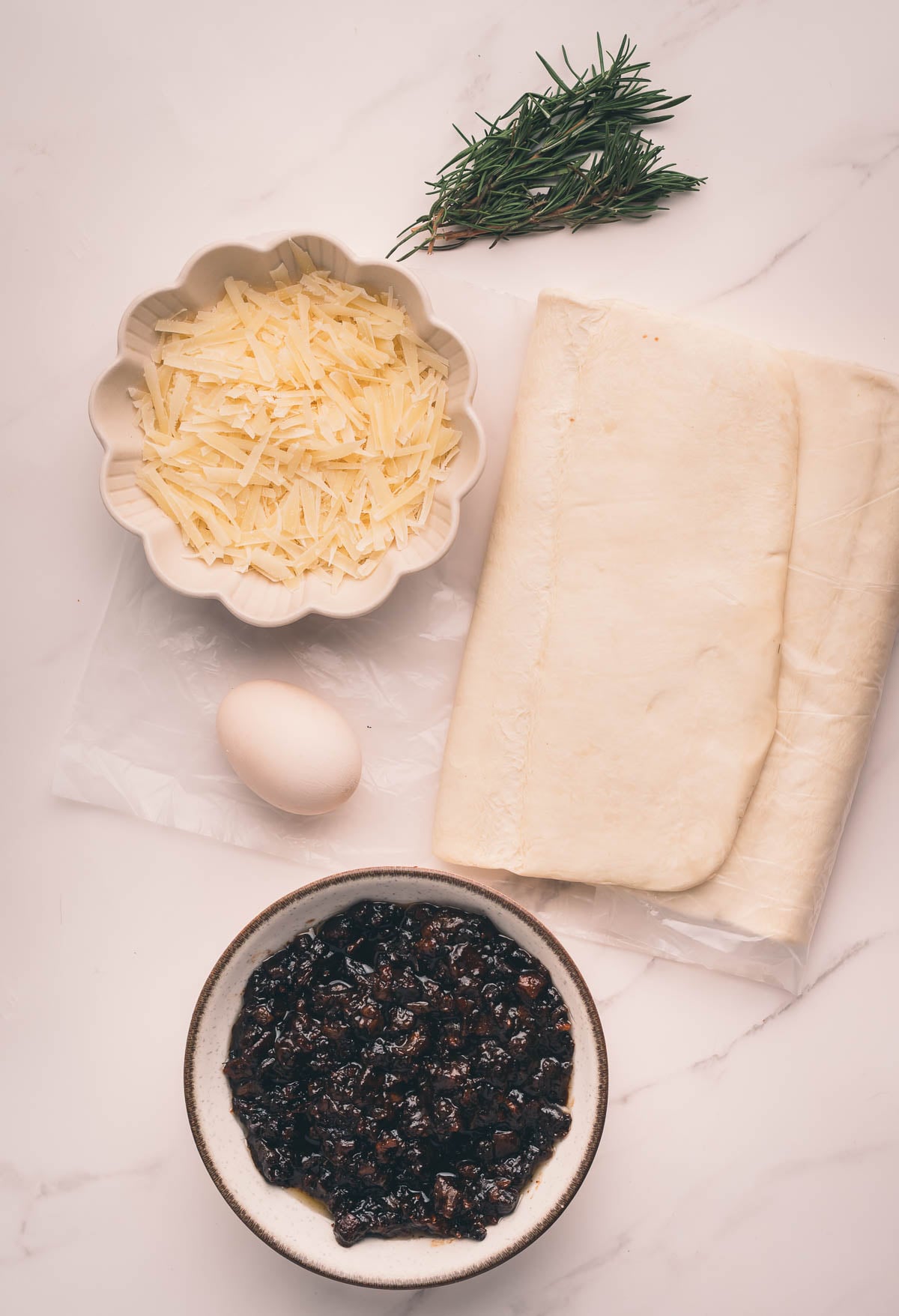 A bowl of grated cheese, a sprig of rosemary, a sheet of puff pastry, a whole egg, and a bowl of bacon jam on a white surface.