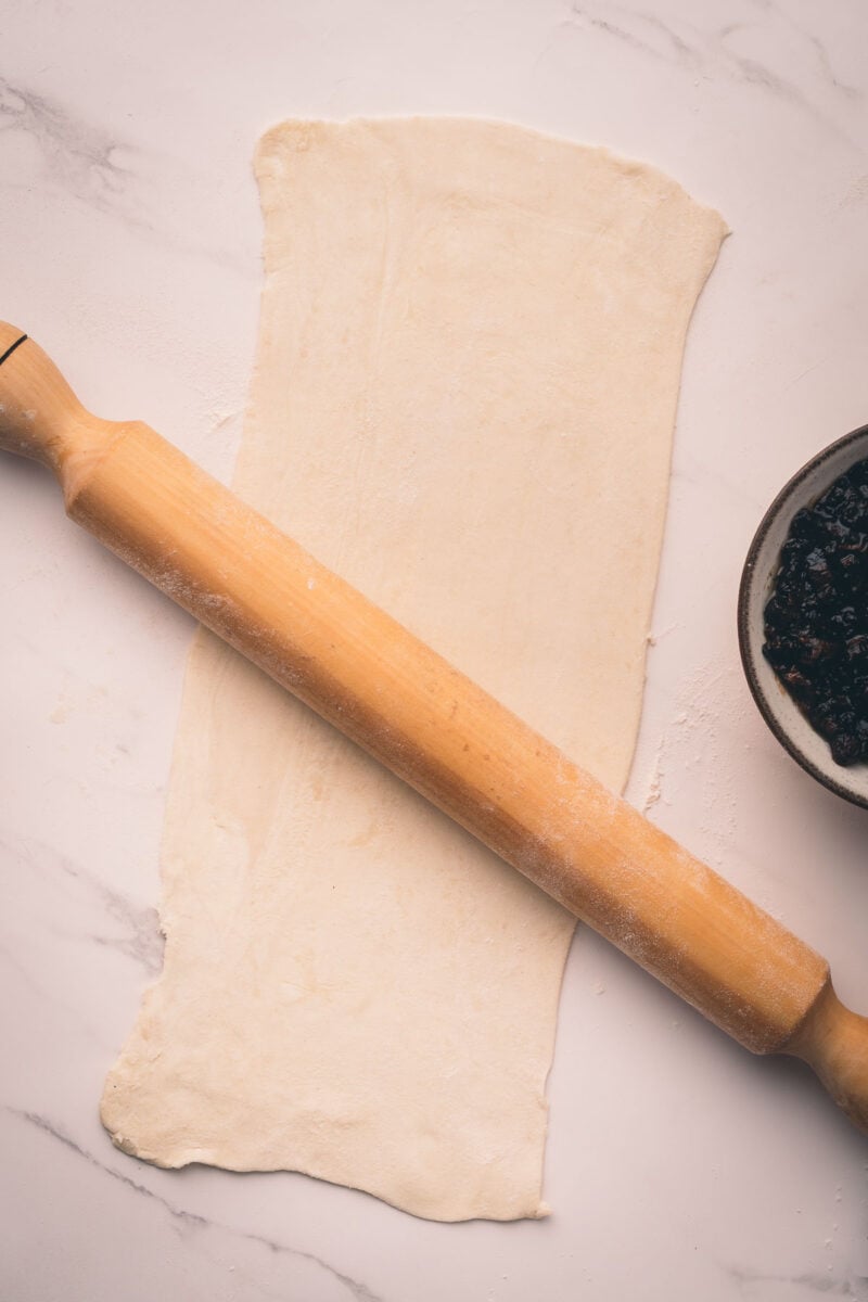 A rolling pin rests on a sheet of rolled-out piff pastry beside a bowl of bacon jam on a marble surface.