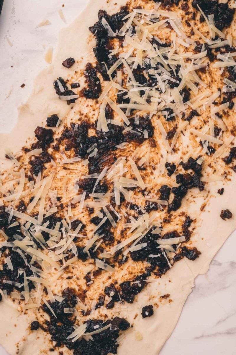A sheet of dough topped with shredded cheese and a dark, chunky bacon jam, laid out on a light marble surface.