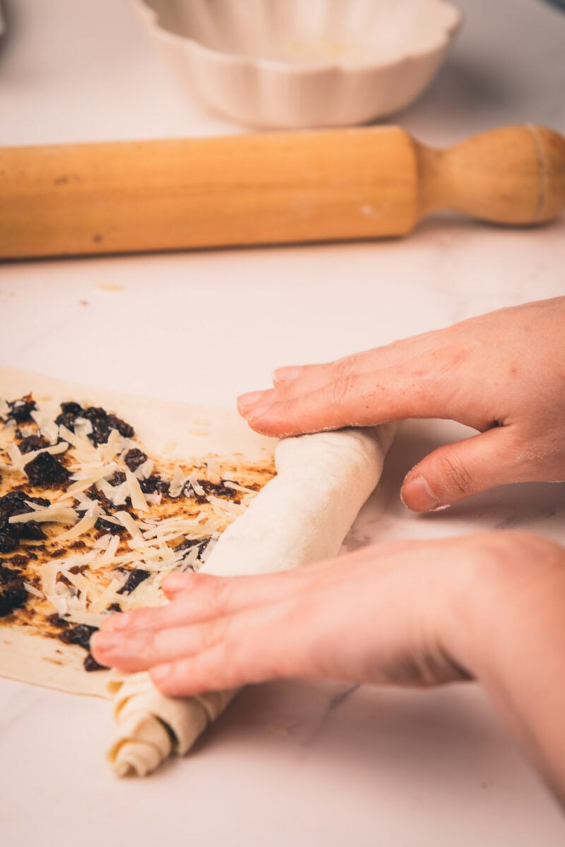 Hands rolling dough filled with grated cheese and bacon jam on a countertop, with a rolling pin and bowl in the background.