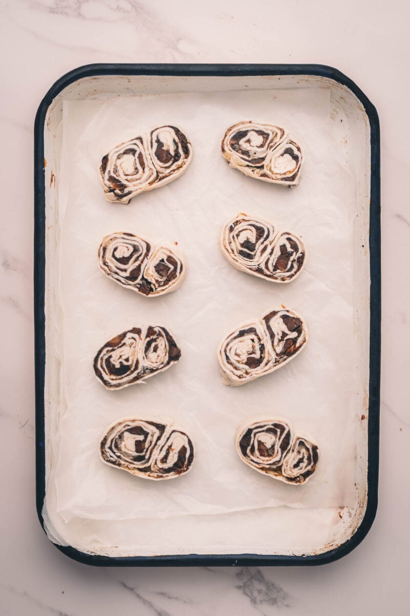 A baking tray lined with parchment paper holds eight unbaked bacon jam palmiers arranged in rows.