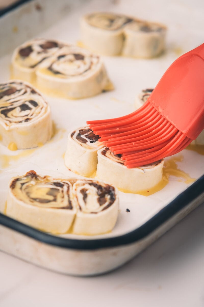 A red silicone brush applies egg wash to unbaked bacon jam palmiers arranged on a parchment-lined baking tray.