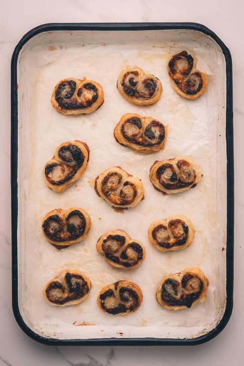 A baking tray with fifteen bacon jam palmier pastries arranged on parchment paper, some with slightly browned edges.