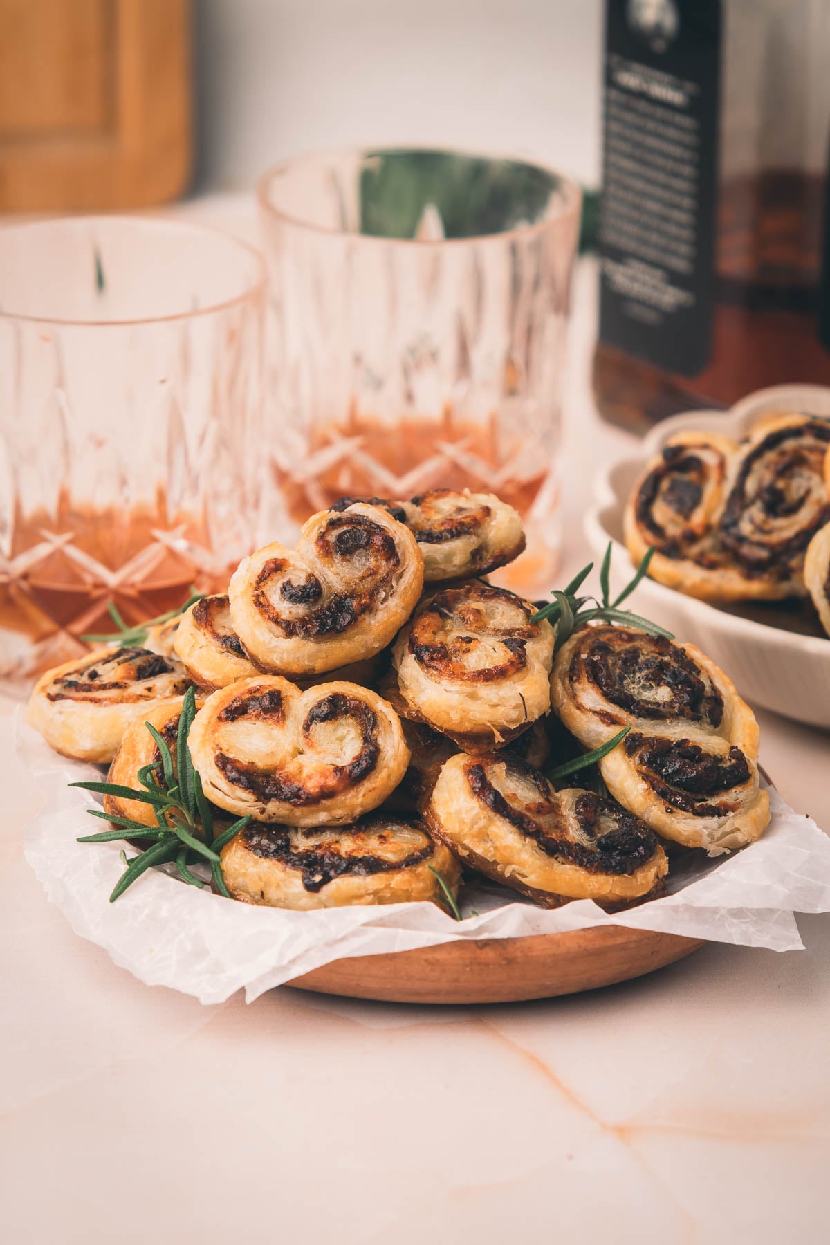 A plate of savory puff pastry pinwheels with bacon jam and herbs, placed on parchment paper, with drinks and more pinwheels in the background.