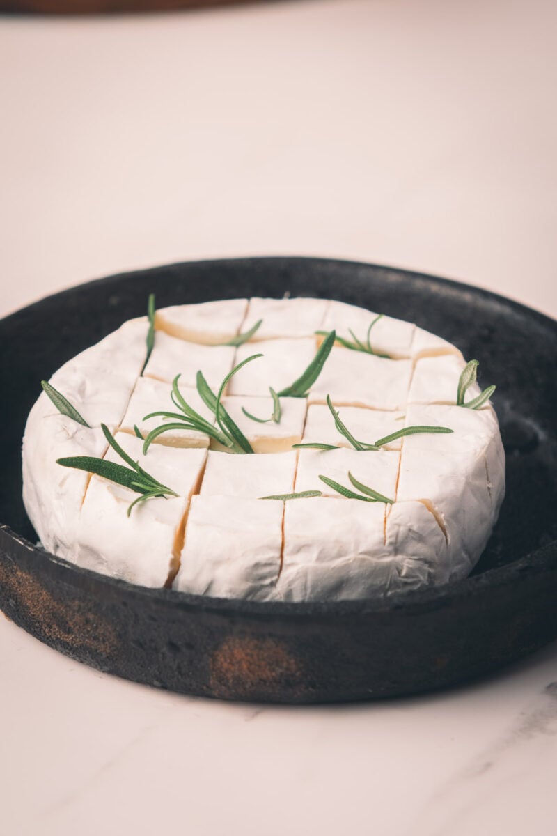 A round wheel of Camembert cheese scored into squares, topped with sprigs of fresh rosemary, sits in a black baking cast iron skillet on a light surface.