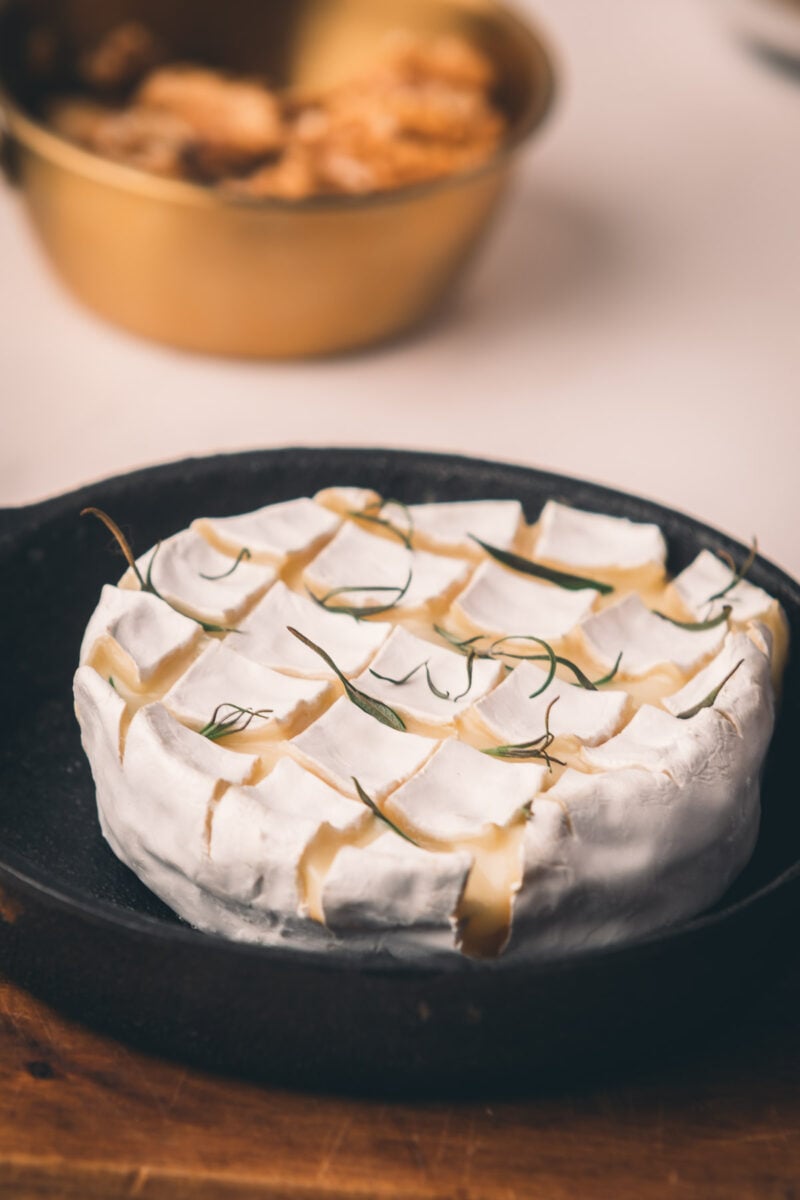 A round wheel of soft baked cheese scored in a crisscross pattern, garnished with herbs, sits in a skillet with a bowl of crackers in the blurred background.