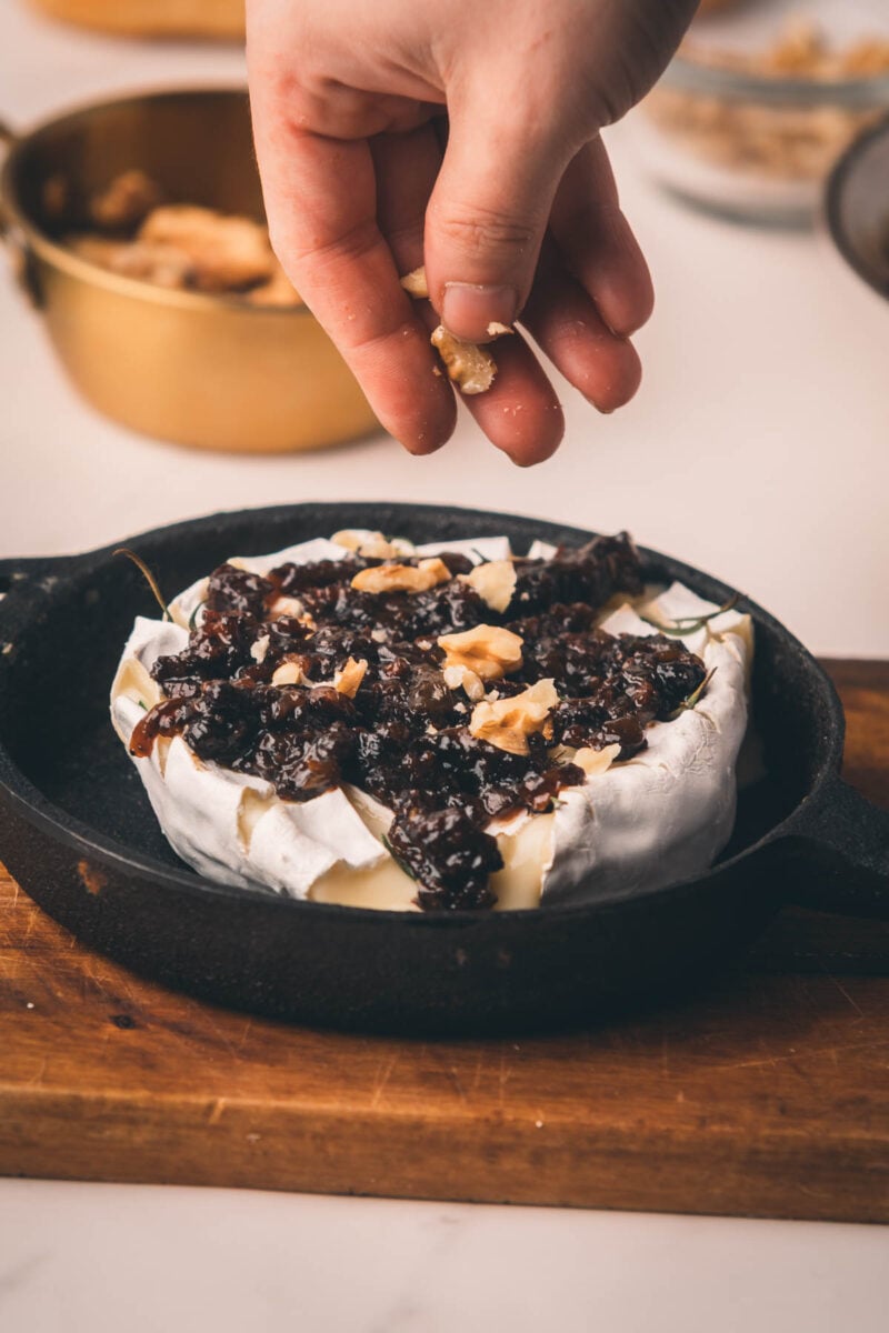 A hand sprinkles chopped walnuts onto a wheel of baked Camembert topped with bacon jam in a small cast iron pan.