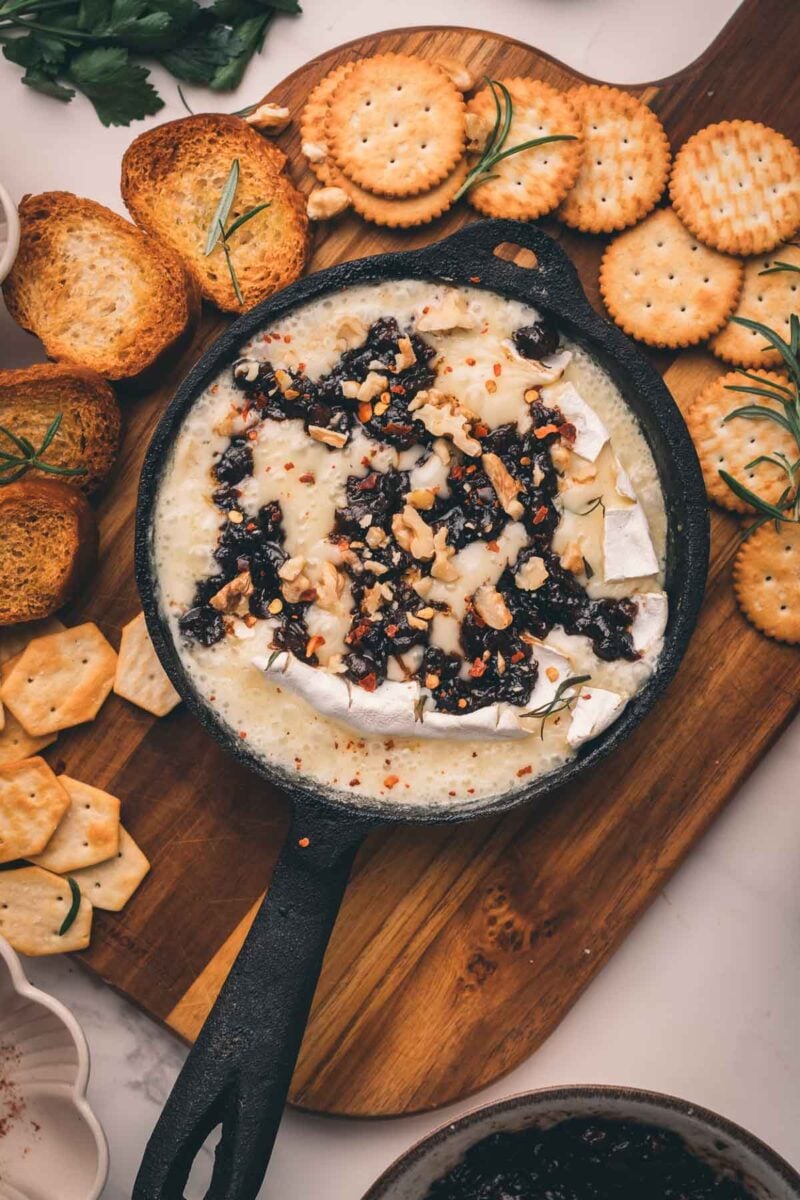 A skillet of melted Camembert cheese topped with chopped nuts and bacon jam, surrounded by crackers and toasted bread on a wooden board.