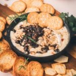 A skillet of baked cheese dip topped with bacon jam and chopped nuts, surrounded by crackers, toasted bread, and fresh rosemary on a wooden board.