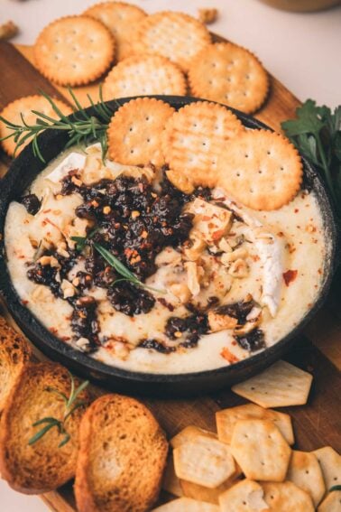 A bowl of baked brie topped with nuts and bacon jam, surrounded by round crackers and toasted bread on a wooden board.